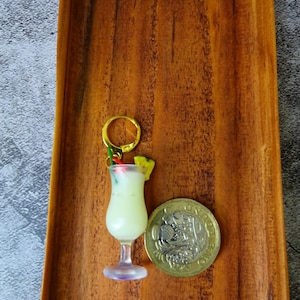 May include: A gold key chain with a miniature plastic cocktail glass filled with a white liquid and topped with a green pineapple slice and a red cherry. The keychain is next to a British one pound coin.