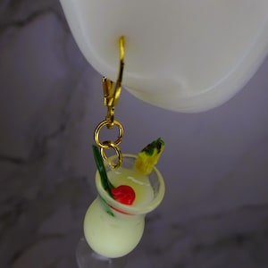 May include: Gold hoop earrings with a yellow and green pineapple and a red cherry on top of a clear glass filled with a white liquid. The drink is a miniature replica of a cocktail.