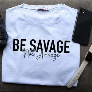 May include: A white t-shirt with the text "BE SAVAGE Not Average" printed in black. The shirt is lying on a brown surface with a camera lens, a black smartphone, and a white Apple Watch.