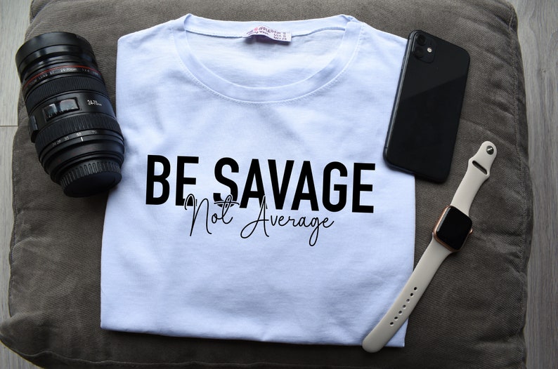 May include: A white t-shirt with the text "BE SAVAGE Not Average" printed in black. The shirt is lying on a brown surface with a camera lens, a black smartphone, and a white Apple Watch.