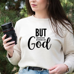 May include: A white sweatshirt with the text "BUT GOD" in black. The text is in a cursive font and is centred on the sweatshirt. The text is surrounded by the smaller text "THERE WAS NO WAY, BUT GOD MADE A WAY."