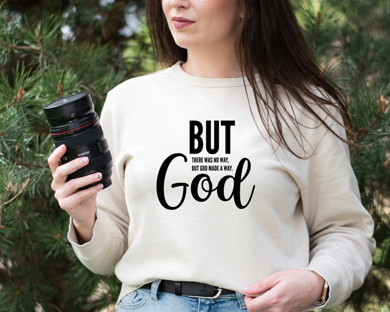May include: A white sweatshirt with the text "BUT GOD" in black. The text is in a cursive font and is centred on the sweatshirt. The text is surrounded by the smaller text "THERE WAS NO WAY, BUT GOD MADE A WAY."