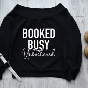May include: Black off-the-shoulder sweatshirt with the text "BOOKED BUSY Unbothered" in white.