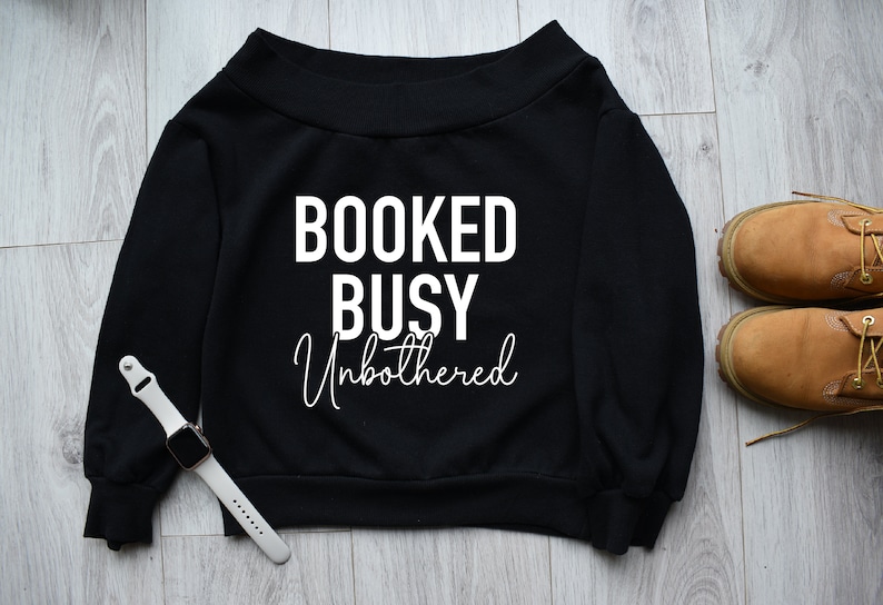 May include: Black off-the-shoulder sweatshirt with the text "BOOKED BUSY Unbothered" in white.