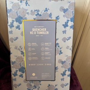 Może przedstawiać: Kartonowe pudełko z kwiatowym wzorem w kolorze jasnoniebieskim i białym. Pudełko zawiera "QUENCHER H2.0 TUMBLER". Pudełko ma złote obramowanie wok&oacute;ł etykiety ze szczeg&oacute;łami produktu. Pudełko ma 1,18 L.