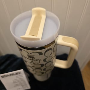 Op de afbeelding: Een witte en beige Stanley reismok met een handvat en een deksel. De mok heeft een patroon van zwarte in elkaar grijpende C-vormen en het Stanley-logo. Een klein wit papier met het Stanley-logo ligt naast de mok.