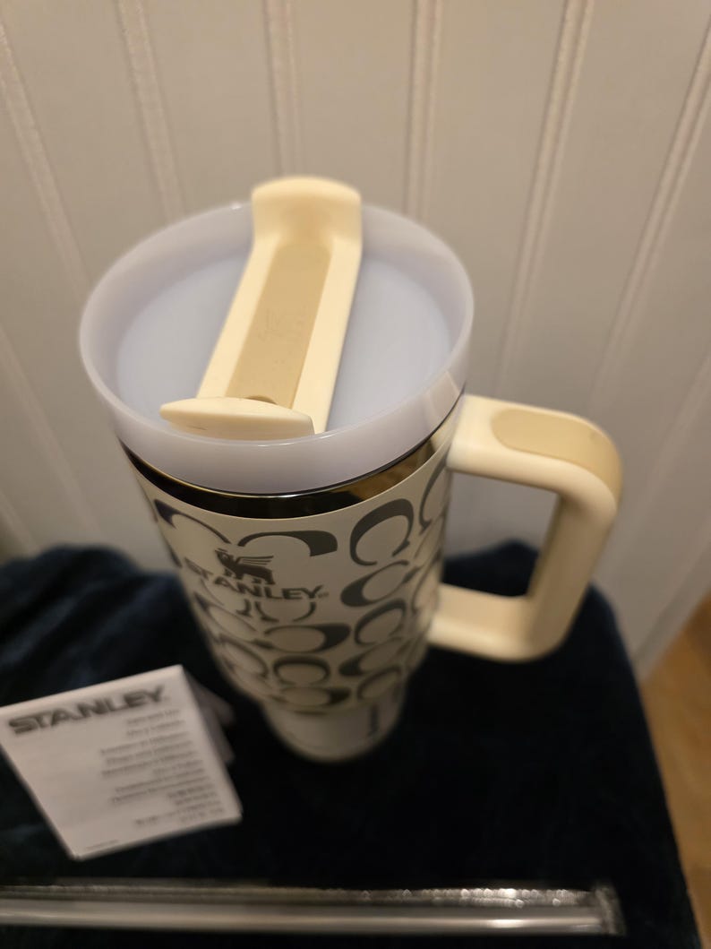 Op de afbeelding: Een witte en beige Stanley reismok met een handvat en een deksel. De mok heeft een patroon van zwarte in elkaar grijpende C-vormen en het Stanley-logo. Een klein wit papier met het Stanley-logo ligt naast de mok.