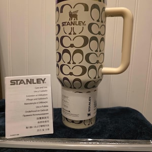 Op de afbeelding: Cr&egrave;mekleurige Stanley reismok met een herhalend patroon van in elkaar grijpende C-vormen. De mok heeft een handvat en een deksel met gouden en iriserende rand. Een verzorgingsboekje en een rietje zijn ook aanwezig.