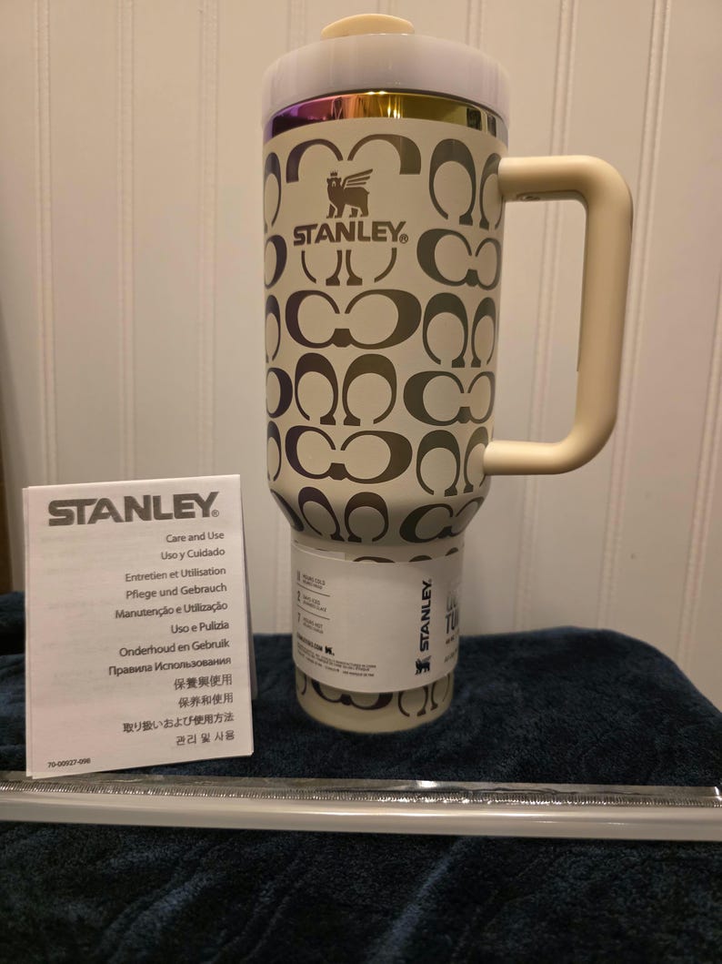 Op de afbeelding: Cr&egrave;mekleurige Stanley reismok met een herhalend patroon van in elkaar grijpende C-vormen. De mok heeft een handvat en een deksel met gouden en iriserende rand. Een verzorgingsboekje en een rietje zijn ook aanwezig.