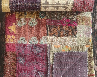 May include: A patchwork quilt with a variety of colors and textures. The quilt features squares of burgundy, olive green, and gold fabrics, some with embroidered floral designs. The quilt is folded over, showing the detailed stitching.