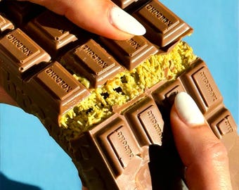 May include: A close-up of a chocolate bar with the word "CHOCOBOX" embossed on each square. The bar is broken open, revealing a light green filling. The background is a solid light blue.