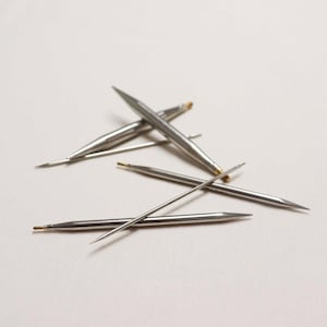 Op de afbeelding: Een set van vijf zilveren breinaalden met puntige uiteinden. De naalden zijn in een kruispatroon op een wit oppervlak gerangschikt.