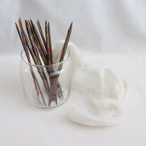Könnte beinhalten: Ein Glasgefäß gefüllt mit einer Sammlung bunter Holzstricknadeln. Die Nadeln sind radial angeordnet, wobei die Spitzen nach außen zeigen. Das Gefäß steht auf einer weißen Oberfläche.
