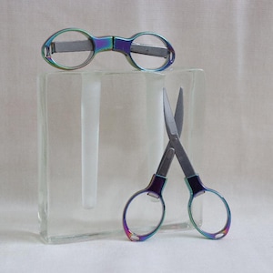 Puede incluir: Un par de tijeras iridiscentes y un par de gafas iridiscentes con una cuchilla plateada. Las tijeras y las gafas están sobre una superficie de vidrio transparente.