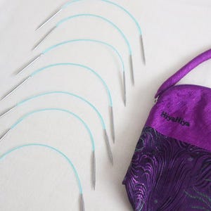 Puede incluir: Un juego de diez agujas de tejer circulares turquesa y plateadas, dispuestas en semicírculo sobre una superficie blanca. Las agujas están en una bolsa con estampado morado y negro con el texto "HiyaHiya" bordado en la parte delantera.