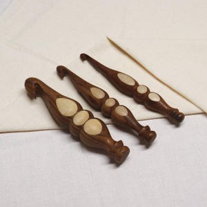 May include: Three wooden crochet hooks with white accents. The hooks are different sizes and are arranged in a row on a white fabric background.