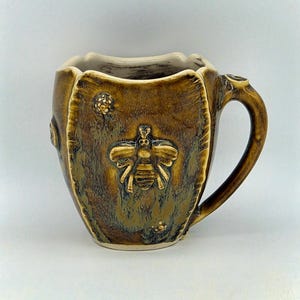 May include: A handcrafted ceramic mug with a bee motif. The mug features a textured, brown glaze with a sculpted bee and honeycomb design. The handle is curved, and the rim has a scalloped edge. The mug is approximately 10 cm tall.