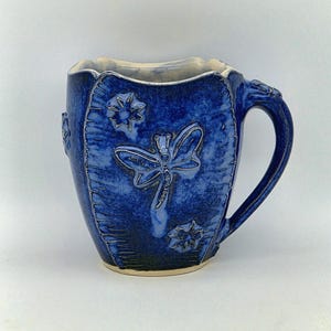May include: A handmade blue ceramic mug with a dragonfly and floral relief design. The mug has a textured surface and a curved handle. The interior is a lighter colour, and the rim has a scalloped edge. A unique piece for enjoying beverages.