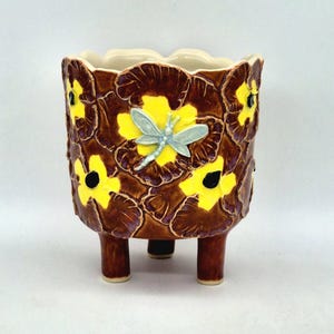 May include: A ceramic planter with a scalloped rim and three short legs. The exterior features a brown glaze decorated with raised yellow pansy flowers with black centres and a single light blue dragonfly relief centred on the front surface.