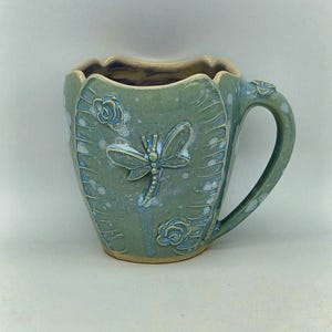 May include: A sage green ceramic mug with a scalloped rim and a curved handle. The exterior features a raised dragonfly relief in the centre, flanked by two small rose motifs. The glaze has a speckled white finish with visible blue undertones.
