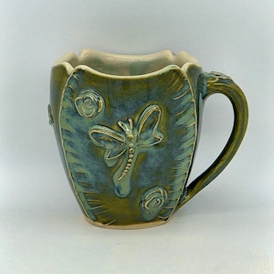 May include: A ceramic mug with a glossy green and blue glaze, featuring a raised dragonfly relief in the centre and small circular floral accents. The mug has a wavy, scalloped rim and a curved handle, with a smooth, light-coloured interior.