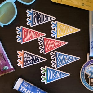 May include: A collection of colorful pennant-shaped stickers featuring the names of cities like Berlin, Boston, and Tokyo, each with "26.2" indicating a marathon distance. The stickers are in various colors, including blue, yellow, and red.