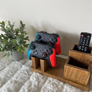 May include: A wooden organiser featuring two black and red video game controllers, a remote control, and two small wooden boxes. A small potted plant is in the background. The controllers have blue and red accents.