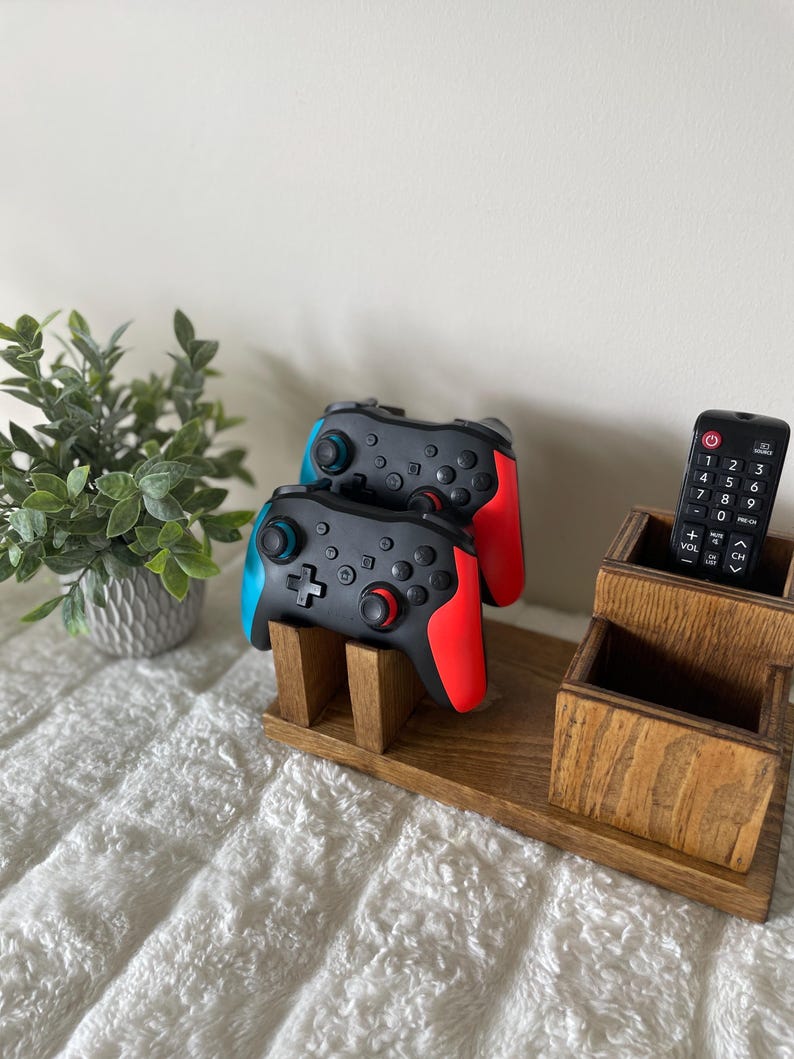 May include: A wooden organiser featuring two black and red video game controllers, a remote control, and two small wooden boxes. A small potted plant is in the background. The controllers have blue and red accents.