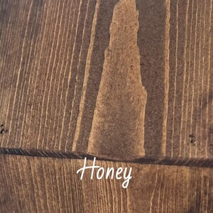 May include: Close-up of a wooden surface with a warm honey-coloured stain. The wood grain is visible, with vertical lines and a lighter, irregular shape in the centre. The word "Honey" is written in cursive.