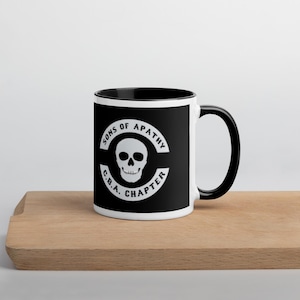 May include: A white ceramic mug with a black interior and handle. The mug features a black square with a white skull and the text "SONS OF APATHY C.B.A. CHAPTER" in a circular design. The mug is on a wooden surface.