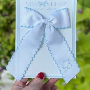 May include: White satin ribbon bow with a light blue scalloped edge and a light blue embroidered letter "B". The bow is on a white card with the text "Winn and William handmade in Dallas, Texas".