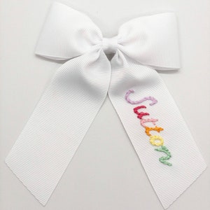 May include: A white grosgrain ribbon bow with long tails. The word "Sutton" is embroidered in colorful thread on one tail. The bow is a classic accessory, suitable for various occasions.