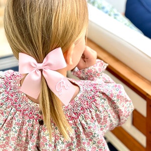 May include: A young girl with blonde hair wearing a pink and white floral dress with a pink bow in her hair. The bow has a white embroidered letter "A" on it.