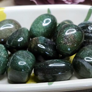 May include: A collection of polished, dark green tumbled stones with subtle black inclusions, contained within a small, rectangular white ceramic dish featuring a painted yellow and green floral pattern along the interior rim and exterior edges.