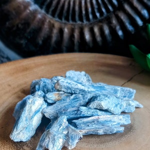 May include: A pile of blue kyanite crystals on a wooden surface. The crystals are long and thin, with a light blue colour and a slightly iridescent sheen.