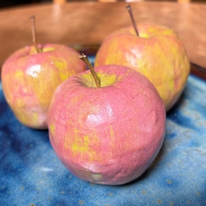 May include: Three decorative apples with a textured, hand-painted finish in shades of pink and yellow. Each apple features a thin, brown stem protruding from the top. The surface of the fruit appears uneven and matte, showing visible brushstrokes.