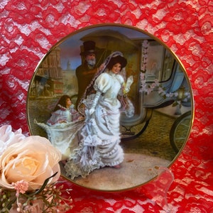 May include: A vintage-style porcelain plate with a gold rim features a painting of a bride in a white dress and veil, a man in a top hat, and a horse-drawn carriage. The scene is set in a city street with buildings in the background.