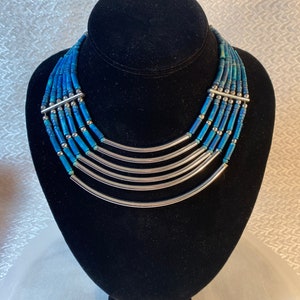 May include: A blue beaded necklace with silver-tone metal accents. The necklace features multiple strands of blue beads, interspersed with silver beads and curved metal bars. The necklace is displayed on a black bust.