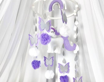 Könnte beinhalten: Ein weißes und violettes Baby-Mobile mit einem Einhorn, Schmetterlingen und Regentropfen. Das Mobile hängt an einem weißen Ring und ist mit weißen Pompons verziert.