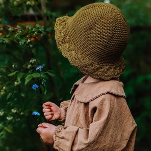 Könnte beinhalten: Ein junges Kind trägt einen braunen, gehäkelten Sonnenhut mit breiter Krempe. Es hält einen Strauß blauer Vergissmeinnicht-Blumen.