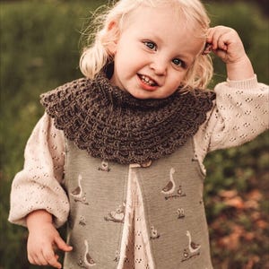 May include: A young girl wearing a brown crocheted cowl and a green vest with a goose pattern. The vest has a button closure and a frayed hem. The girl is wearing a white dress with a lace pattern.