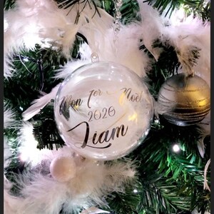 Peut inclure: Une boule de Noël en verre transparente avec des plumes blanches à l'intérieur. La boule est ornée d'une inscription dorée "Mon 1er Noël 2020 Liam".