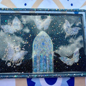 May include: A black resin tray with a glittery finish features a stained glass window design and five white butterflies. The tray is perfect for holding jewelry, keys, or other small items.