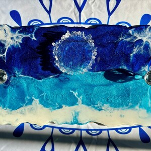 May include: A blue and white resin tray with a wavy design, resembling ocean waves. The tray has a clear crystal-like circle in the center and two clear crystal-like accents on the sides.
