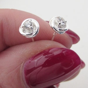 May include: A pair of silver stud earrings with a small clear rhinestone in the center of each earring. The earrings are shaped like a rose.