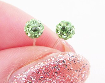 Op de afbeelding: Een paar kleine, groene, glimmende oorbelletjes. De oorbelletjes worden vastgehouden in een hand met roze nagellak.