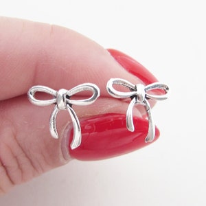 May include: A pair of small, silver bow-shaped stud earrings. The earrings are held between fingers with red nail varnish. The bows have a detailed design, with the loops and tails clearly defined. These earrings are a delicate accessory.