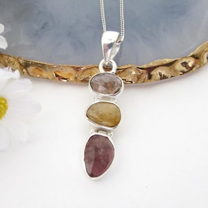 Può includere: Una collana con pendente in argento caratterizzata da tre pietre preziose sfaccettate impilate verticalmente nei toni del marrone chiaro, giallo dorato e rosa scuro. Le pietre sono incastonate in argento lucido su una catenina sottile.