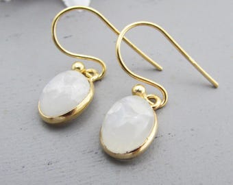 May include: A pair of gold-colored drop earrings with oval, faceted, white gemstones. The earrings have a simple, elegant design with a gold bezel setting and a hook closure. The background is a soft gray.