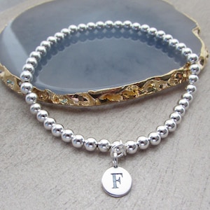 Könnte beinhalten: Ein silbernes Perlenarmband mit einem runden Anhänger. Der Anhänger trägt den eingravierten Buchstaben "F". Das Armband besteht aus kleinen, runden, glänzenden Perlen. Das Armband wird auf einer grauen Oberfläche präsentiert.